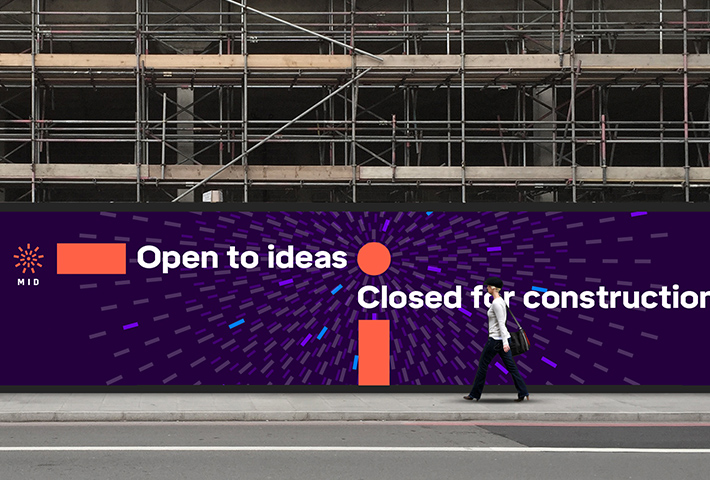 A person walks in front of construction hoarding with MID graphics and the tagline "Open to ideas, closed for construction".