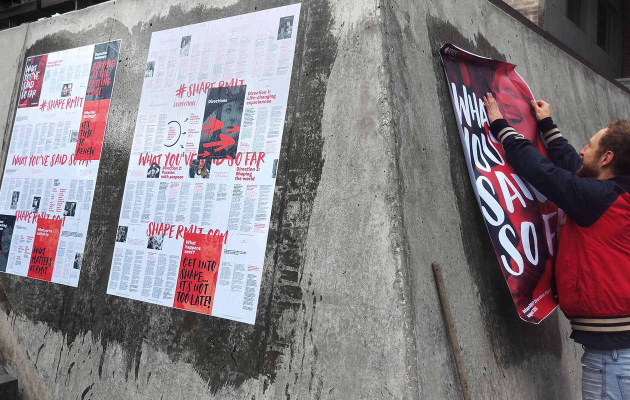An installer putting up three wheatpaste posters in the RMIT city campus. The poster content is a compilation of staff and student responses from the ShapeRMIT campaign.