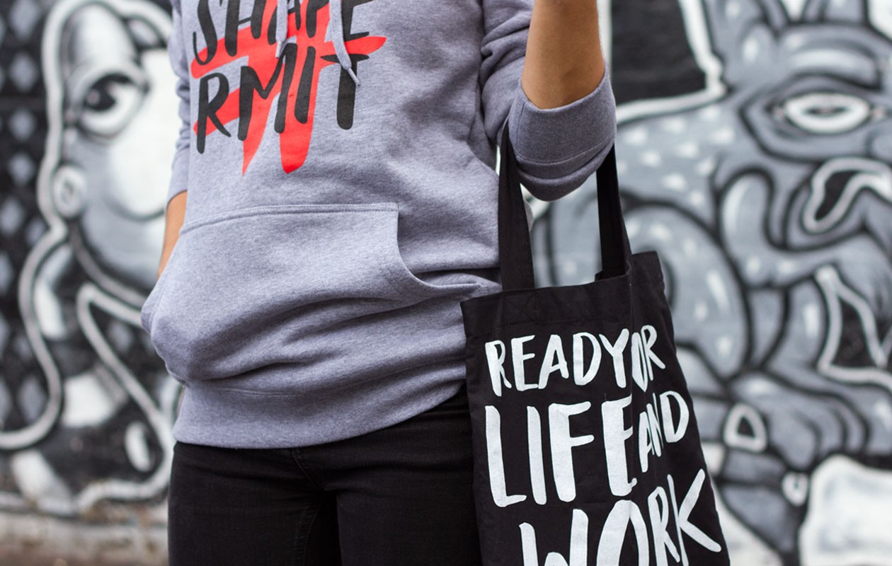 Person wearing grey hoodie with the ShapeRMIT brand mark printed on the front, holding a black tote bag with the words "Ready for Life and Work".