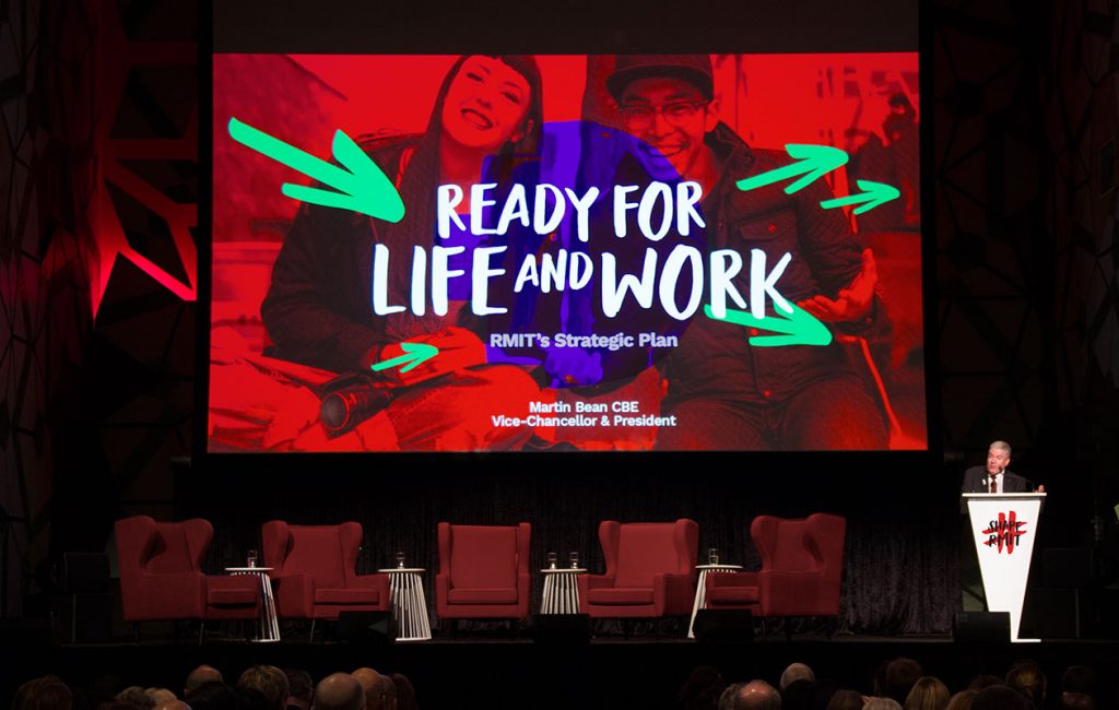 ShapeRMIT Launch keynote presentation. A large screen with a image with the heading "Ready for Life and Work", and a speaker standing at a podium to the lower right hand side of the screen.