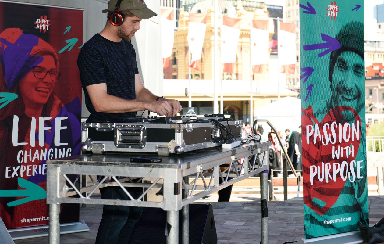 A music DJ standing at turntables in an outdoor setting flanked by two ShapeRMIT campaign pull up banners with the tag lines "Life Changing Experiences" and "Passion with Purpose" respectively.